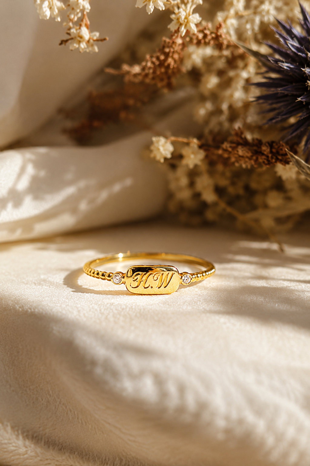 A gold name ring with the name 'JON' engraved on a polished plate, flanked by small clear stones, resting on draped cream fabric with dried flowers in the background.